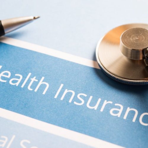 pen and stetoscope on top of of a piece of paper that says "health insurance" in white text over a blue background