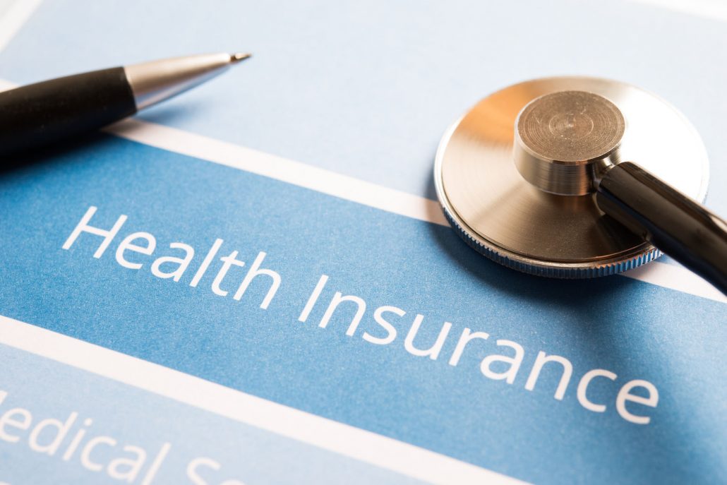 pen and stetoscope on top of of a piece of paper that says "health insurance" in white text over a blue background