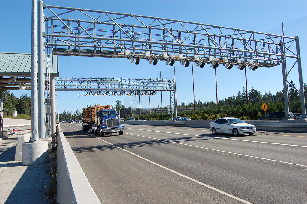 lumber truck and white car passing under highway toll gantry