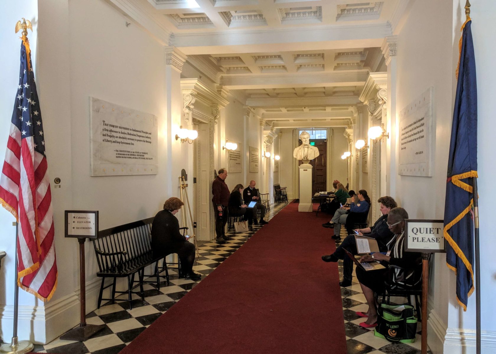 people waiting in hallway with maroon carpet on checkered floor