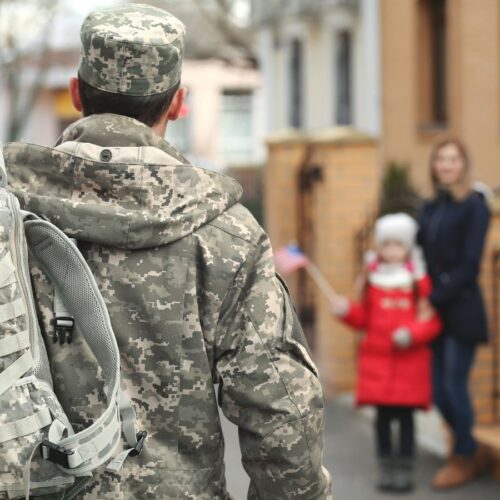 Man in military-issued camo walking away from camera, towards out-of-focus mother and child.
